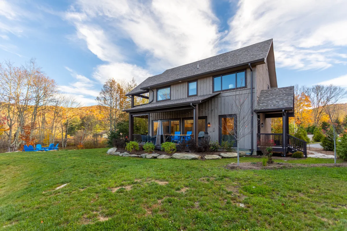 This exterior image features the house set against a backdrop of mature trees and a spacious, grassy yard. Blue chairs invite relaxation on the porch, while the open outdoor space hints at possibilities for games, gatherings, or simply soaking in the mountain air. The overall scene captures the spirit of outdoor adventure and connection with nature, while the porch and fire pit area suggest year-round enjoyment.
