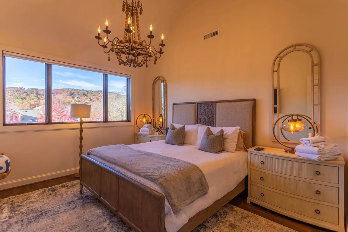 A serene king suite bedroom featuring a plush bed set beneath a chandelier, flanked by a nightstand and decorative mirror. Large windows flood the space with natural light and offer tranquil views, while the soft furnishings and tasteful decor create an atmosphere of quiet luxury and relaxation.