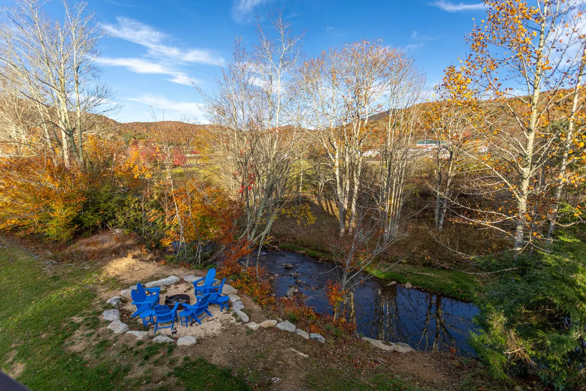 A cozy outdoor fire pit area surrounded by blue chairs, set beside a gently flowing stream and framed by vibrant autumn foliage. The setting is inviting for stargazing, storytelling, or simply unwinding by the fire.