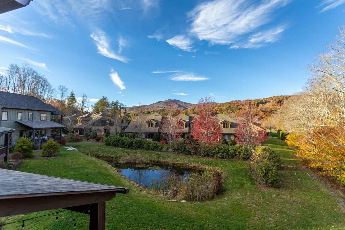 A sweeping view of the mountain landscape with a tranquil pond in the foreground, vibrant foliage framing the scene, and tasteful residential buildings nestled at the edge. The scene evokes the serene natural beauty that surrounds Maynard Rocks IV, with hints of walking paths and quiet spots for reflection or romantic strolls.