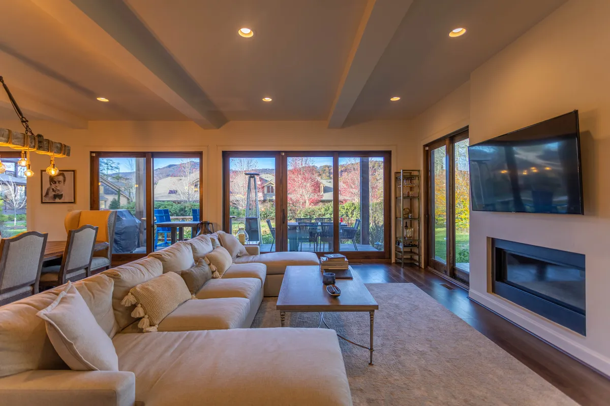 A spacious living area with a plush sectional sofa faces a wall-mounted smart TV, with expansive windows that let in abundant natural light and frame picturesque outdoor views. A coffee table and cozy details create an inviting space ideal for relaxing, entertaining, or movie nights by the fireplace (not in the frame but referenced in the mood).