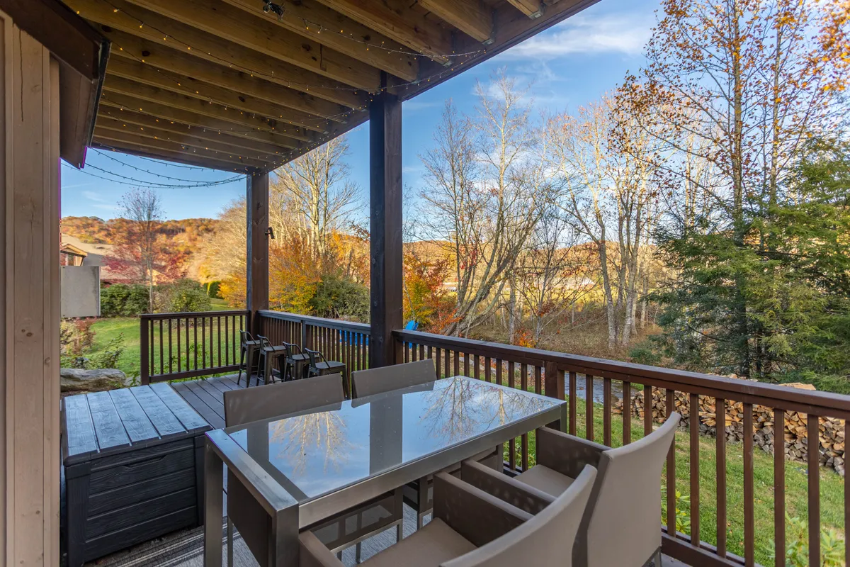 A wooden deck surrounded by lush greenery, featuring a spacious dining table set for outdoor meals. The deck overlooks a serene landscape with tall trees and offers a welcoming spot to enjoy nature throughout the year.