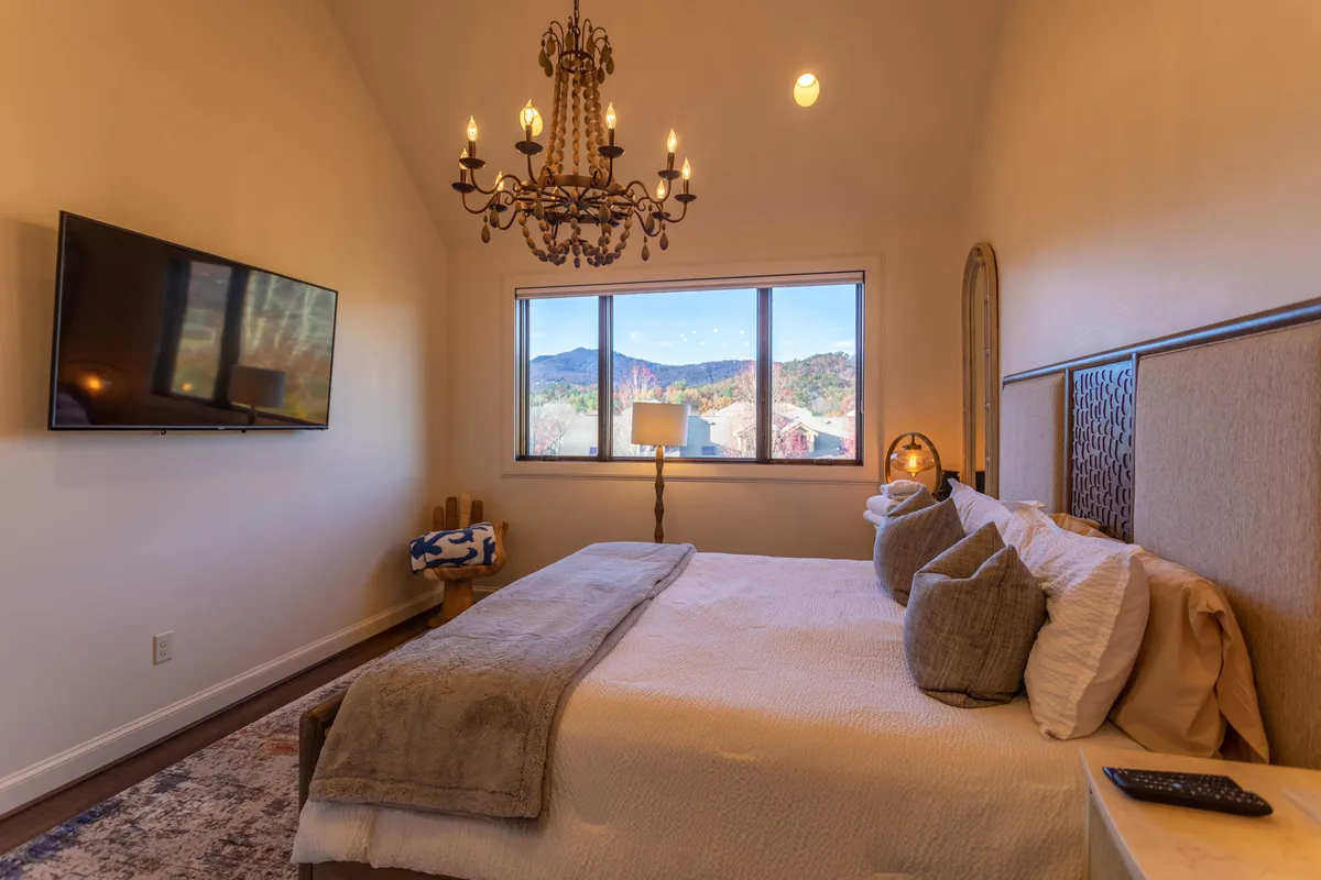 Another king suite bedroom with a comfortable bed, elegant chandelier overhead, large window showcasing mountain vistas, and a wall-mounted smart TV. The space is designed with soothing tones and uncluttered style, reinforcing a restful and upscale ambiance.