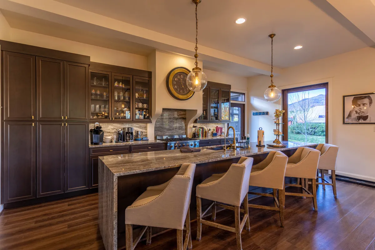 This image showcases the modern chef’s kitchen, featuring a sleek island with bar stools, elegant cabinetry, pendant lighting, and a window that offers a view outside. The scene is styled for entertaining and cooking, with a warm, welcoming ambiance that encourages gatherings and culinary creativity.