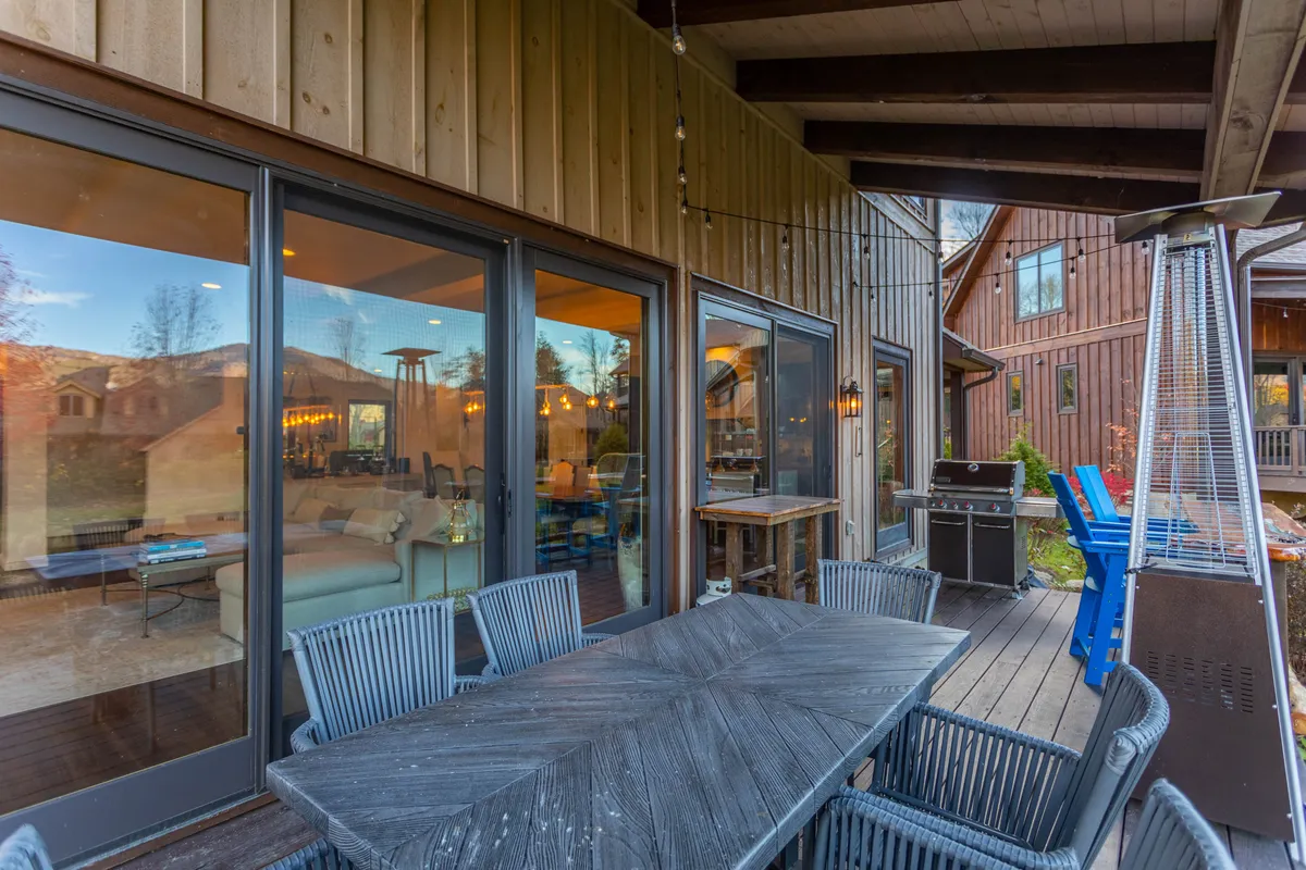 An exterior scene showcasing a stylish outdoor dining table with heaters, a grill, and large glass doors opening into the home. The surrounding landscape, visible in the background, enhances the sense of being immersed in nature. The setup is ideal for group meals and socializing, emphasizing both adventure and comfort.