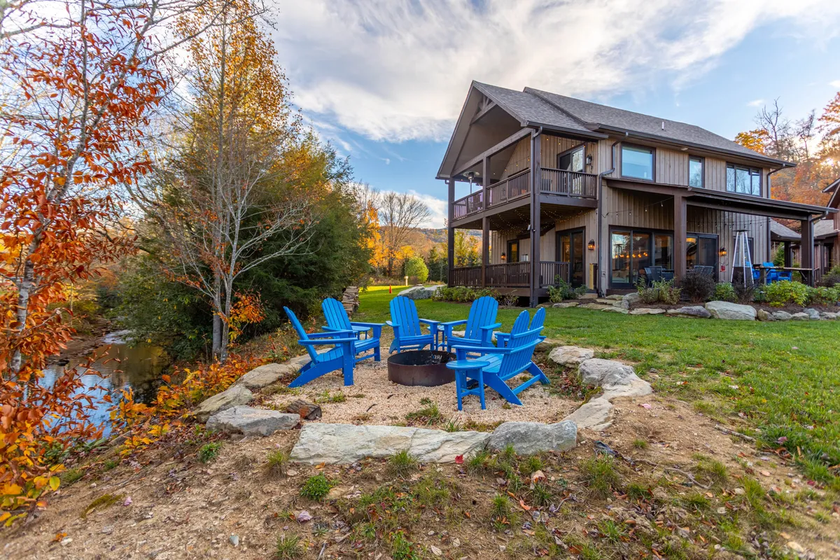 The hero image showcases a charming two-story house nestled in a landscaped garden, with inviting blue Adirondack chairs gathered around a glowing fire pit. The outdoor space feels cozy and welcoming, perfect for relaxing, sharing stories, and enjoying the tranquil natural setting just outside Banner Elk.